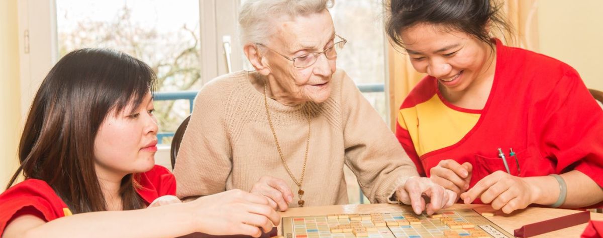 Generalistische Pflegeausbildung – Azubis der Vivantes Hauptstadtpflege im Interview: Das Bild zeigt zwei weibliche Pflegekräfte aus Vietnam und in der Mitte eine Seniorin. Zwei sitzen, die Pflegekraft links hockt am Tisch und spielen Scrabble.