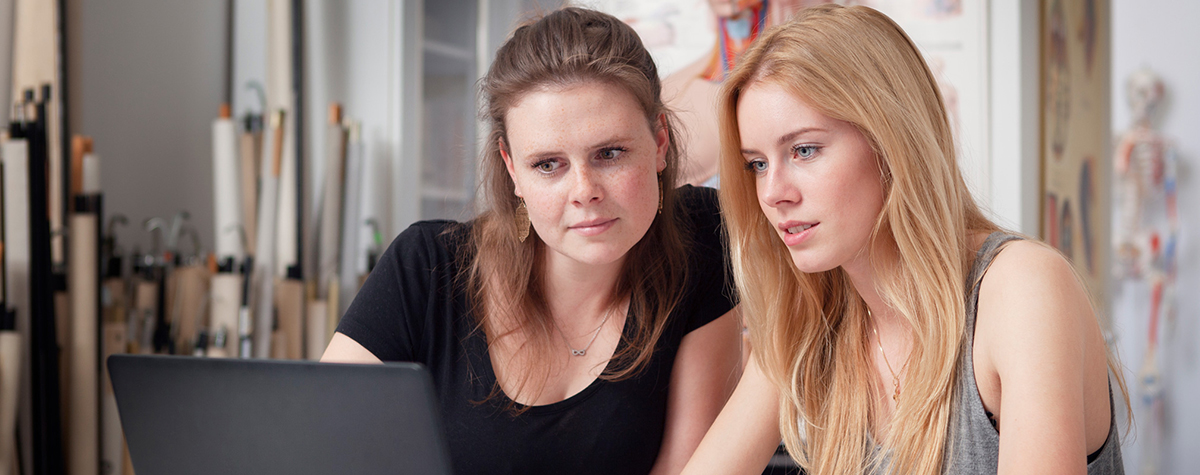 Zwei junge Frauen schauen auf einen Laptop, im Hintergrund sieht man das Anatomie-Labor