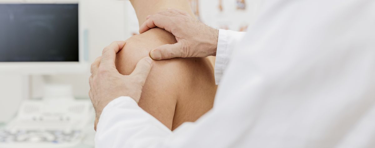 Schulter: Schmerzen, Verletzungen und Erkrankungen – welche gibt es?: Das Bild zeigt den linken Arm eines Arztes in weißem Kittel von der Seite, der mit seinen Händen die linke Schulter eines Patienten untersucht.