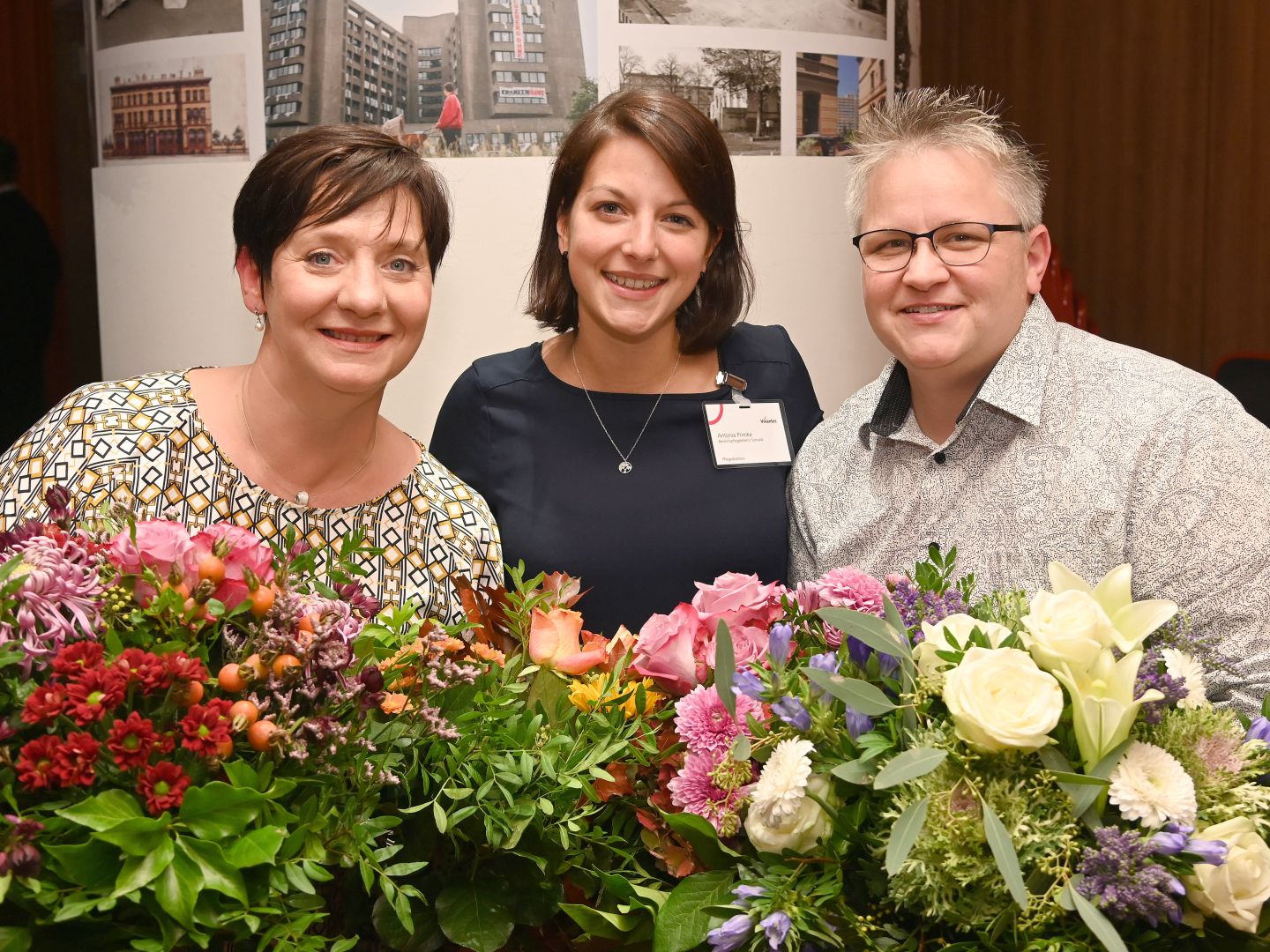 Pflege im Krankenhaus in Berlin-Kreuzberg: Links ist die ehemalige, rechts die neue Pflegedirektorin und zwischen ihnen eine Bereichspflegeleiterin posieren lächelnd mit Blumensträußen während der Amtsübergabe.