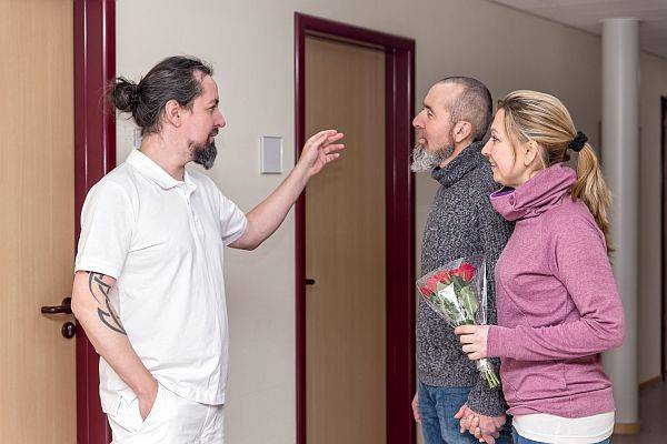 Pfleger zeigt Paar mit Blumen den Weg zur neurologischen Station