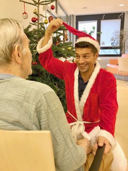 Pflege bei Demenzerkrankungen in der Psychiatrie in Berlin-Kreuzberg: Das Bild zeigt einen Pfleger im Weihnachtsmannkostüm, der mit seiner rechten Hand seine Zipfelmütze nach oben zieht und vor einem Stuhl hockt, in dem ein betagter Herr sitzt.