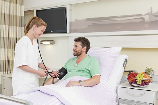Aufgaben der Pflege im Krankenhaus Neukölln: Das Bild zeigt eine Pflegerin, die den Blutdruck einen im Bett liegenden Patienten misst.