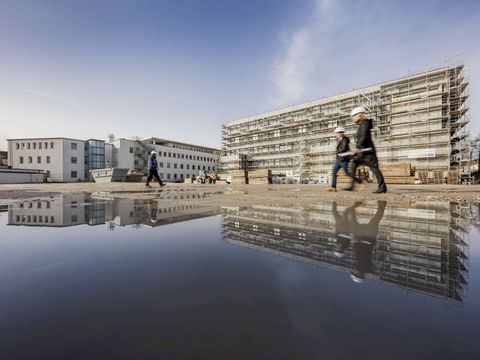 Pflege im Krankenhaus in Berlin-Schöneberg: Vor noch unfertigen Neubauten, die sich im Wasser spiegeln, laufen drei Personen mit Schutzhelm über die Baustelle.. 