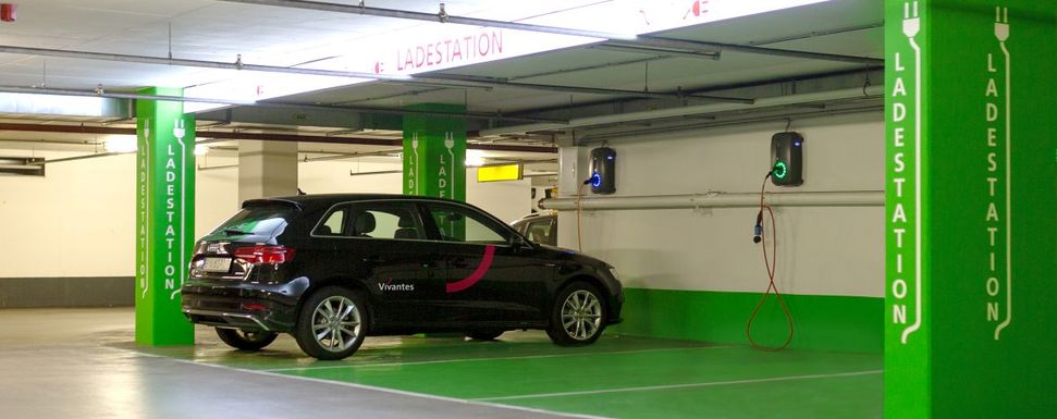 Vivantes for Future – Energiesparen im Krankenhausunternehmen: Das Bild zeigt drei Parkplätze mit je einer Ladestation für Elektroautos in einer Tiefgarage. Auf dem Parkplatz links steht ein schwarzes Auto mit dem Logo von Vivantes, das aufgeladen wird.