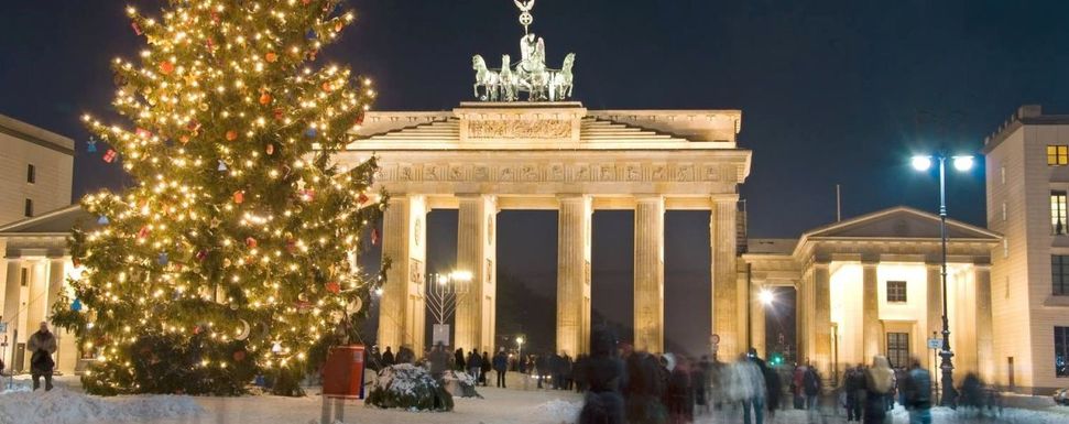 Corona-Weihnachten feiern – Tipps fürs Fest: Das Bild zeigt das beleuchtete Brandenburger Tor in Berlin an einem Dezemberabend. Links ist ein großer geschmückter und beleuchteter Weihnachtsbaum zu sehen. Personen sind nur schemenhaft erkennbar.