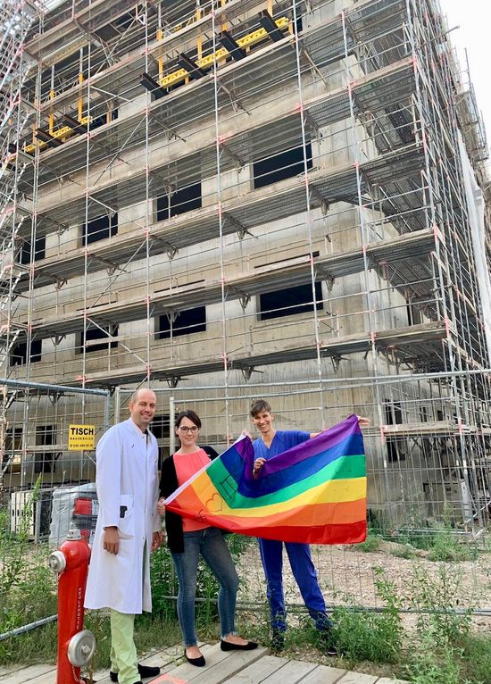 Neubau Auguste-Viktoria-Klinikum in Berlin-Schöneberg: Drei Mitarbeitende, darunter die Pflegedirektorin, posieren vor einem Rohbau mit Regenbogenfahne.