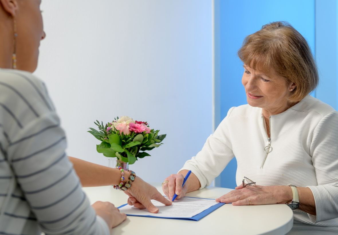 Frau und ältere Patientin sitzen an einem runden weißen Tisch mit kleinem Blumenstrauß drauf und Frau zeigt Patientin einen Bogen, den sie ausfüllen muss