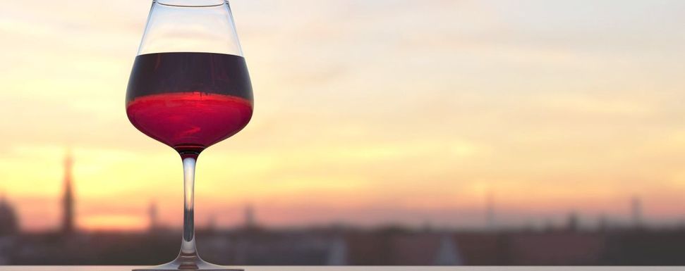 Alkoholsucht in der Familie – Tipps im Umgang mit Kindern: Das Bild zeigt links ein Weinglas, das mit Rotwein gefüllt ist und auf einer Brüstung steht, sowie verschwommen im Hintergrund die Skyline einer Stadt bei Dämmerung.