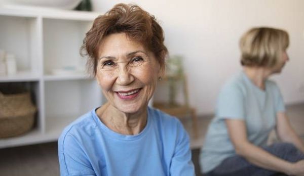 ältere Dame in hellblauen Shirt lächelt in die Kamera, Hintergrund weitere Frau mit blonden Haaren