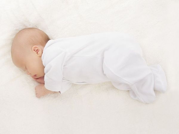 Schlafen wie ein Baby: Das Bild zeigt einen Säugling in einem weißen Strampler. Das Baby befindet sich in Seitlage auf einer cremefarbenen Decke, die rechte Hand liegt teils unter dem Kopf.