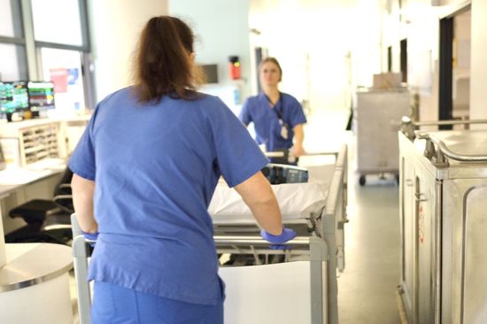 Pflege im Krankenhaus Neukölln: Das Bild zeigt zwei Pflegerinnen, die eine Krankenliege mit Rollen durch einen Krankenhausflur bewegen