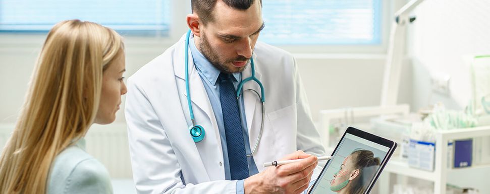 Arzt erklärt einer Patientin mithilfe eines Stiften und einem Tablet-Gerät. Auf dem Tablet sieht man ein Gesicht. Im Hintergrund ist ein Besprechungszimmer sichtbar.