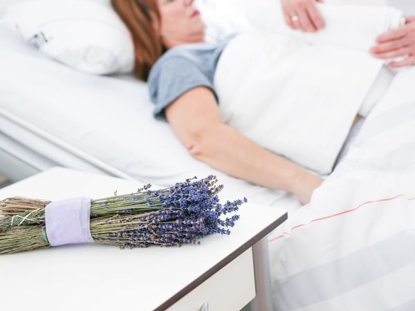 Heilende Düfte – Aromatherapie in der Altersmedizin: Das Bild zeigt im Vordergrund links ein Bündel Lavendel auf einem Nachttisch und im Hintergrund unscharf die obere Hälfte eines Krankenhausbettes, in dem eine Patientin liegt. Oben rechts sind zwei Hände zu sehen, die bei der Patientin einen Bauchwickel anlegen.