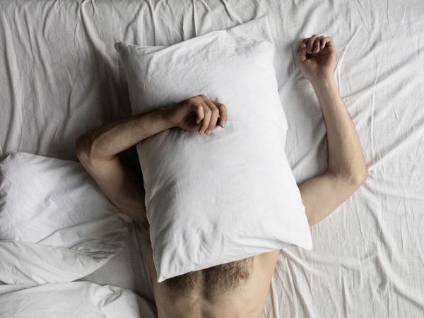 Grübeln raubt den Schlaf: Das Bild zeigt den Oberkörper eines Mannes, der im Bett liegt und sich ein Kopfkissen über seinen Kopf-, Hals- und Brustbereich gelegt hat, wobei die rechte Hand auf dem Kissen ruht. 
