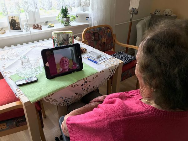 Auf dem Bild ist eine Bewohnerin aus der Schulterperspektive zu sehen, vor ihr steht auf dem Tisch ein iPad, auf welchem eine Videokonferenz läuft