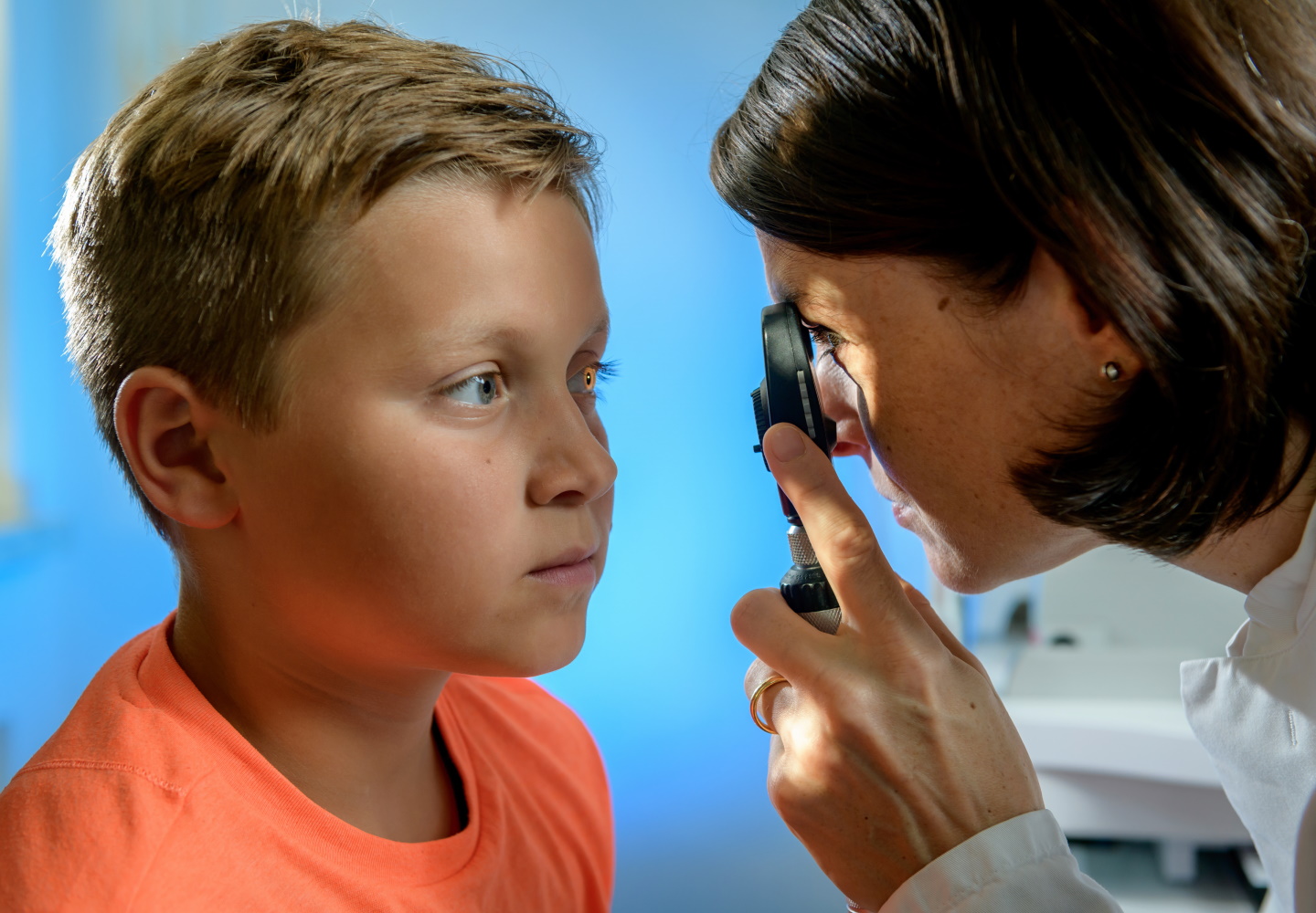 Junge mit blonden kurzen Haaren und orangenem Shirt wird an seinem Auge von Ärztin mit Lupe untersucht